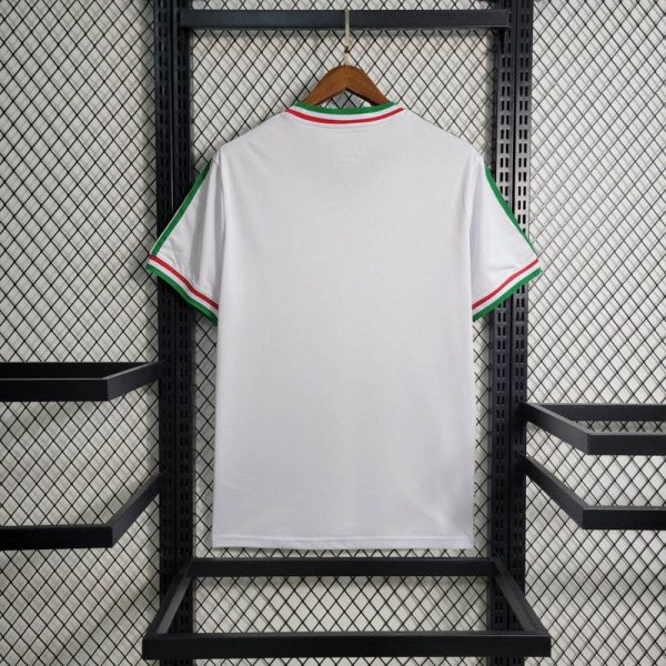 White T-shirt with green, white, and red trim on collar and sleeves, hanging on a wooden hanger against a black metal rack (back view).