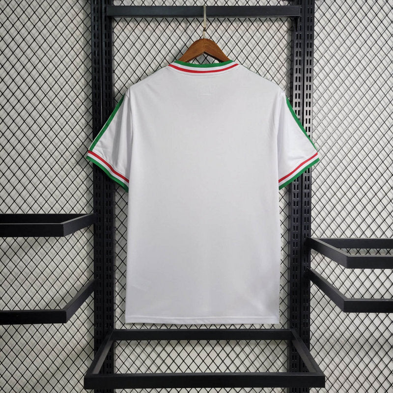 big-620_5e034418-36d5-42e0-92b8-9536753c1b36 | UAE SPORT STORE White T-shirt with green, white, and red trim on collar and sleeves, hanging on a wooden hanger against a black metal rack (back view).