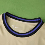 Beige athletic jersey collar with black and purple trim, logo visible inside neck area; green background behind the shirt