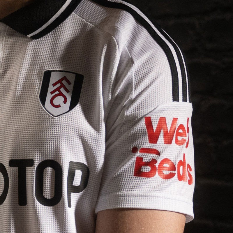 bm50M4N6NvUVXg7 | UAE SPORT STORE Close-up of a white Fulham FC jersey showing the crest on the left chest and red sponsor text on the sleeve.