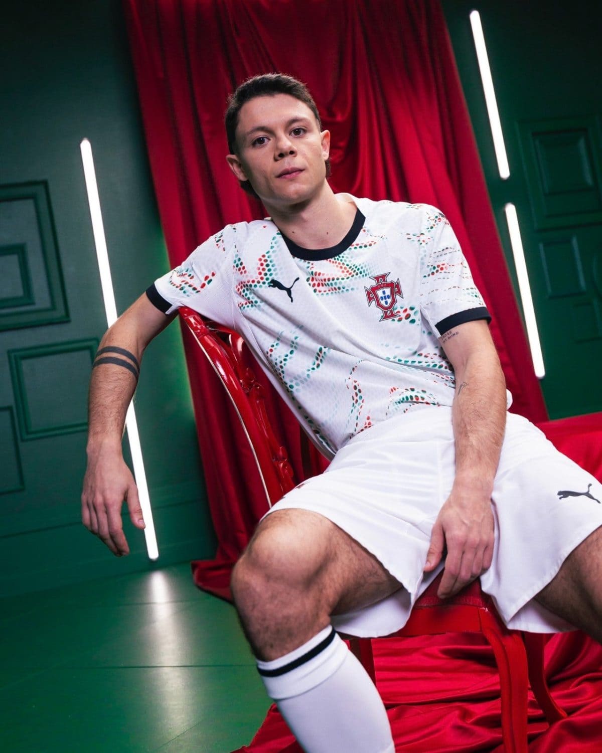 Young man in a Portugal national team jersey sits on a red chair, with a red curtain and green walls in a neon-lit studio.