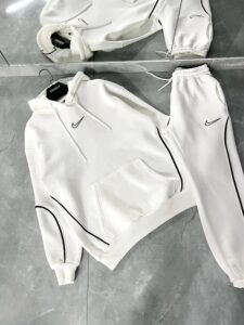 White Nike tracksuit laid flat on a gray tiled floor, hoodie and joggers with black piping and logos.