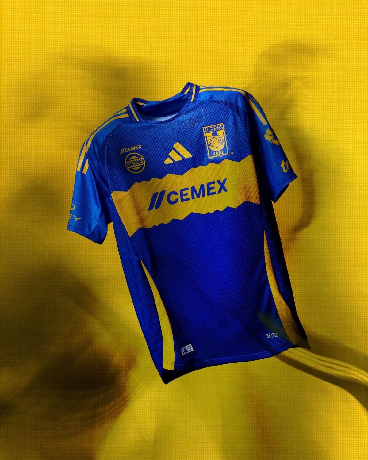 Blue and yellow soccer jersey with CEMEX sponsor and team crest, suspended against a yellow backdrop of the studio light.