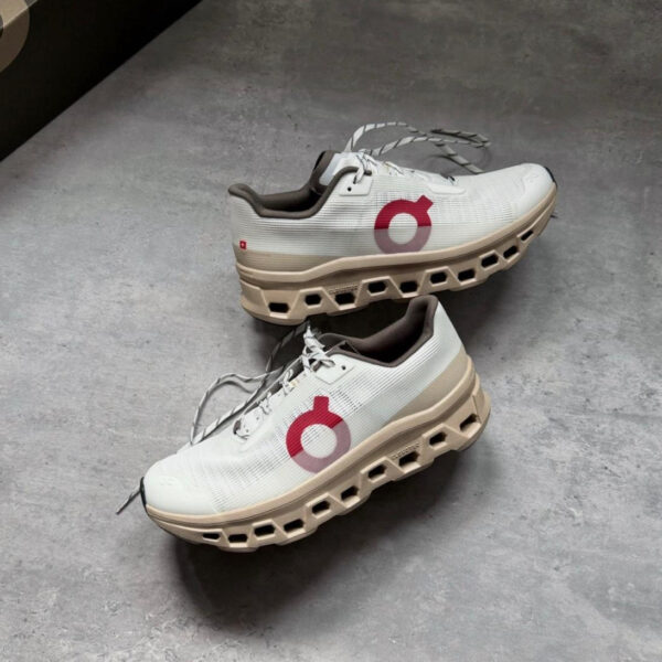 Pair of white and beige running shoes with a chunky, hollowed-out beige sole and red circular logo on the side, lying on a gray floor; laces loose.