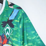Close-up of a green camouflage jersey with a green diamond crest displaying a white stylized 'W' logo on the chest side.