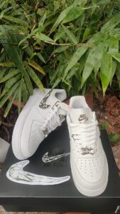 Pair of white Nike Air Force 1 sneakers with metal lace charms, resting on a black box beside green plants.