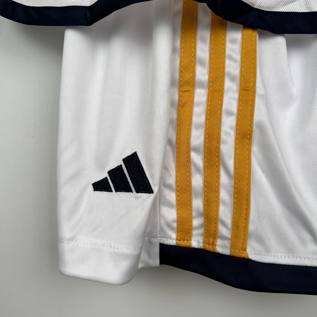 Close-up of white fabric showing three orange striped straps stitched down, with part of a black Adidas logo in the bottom left.