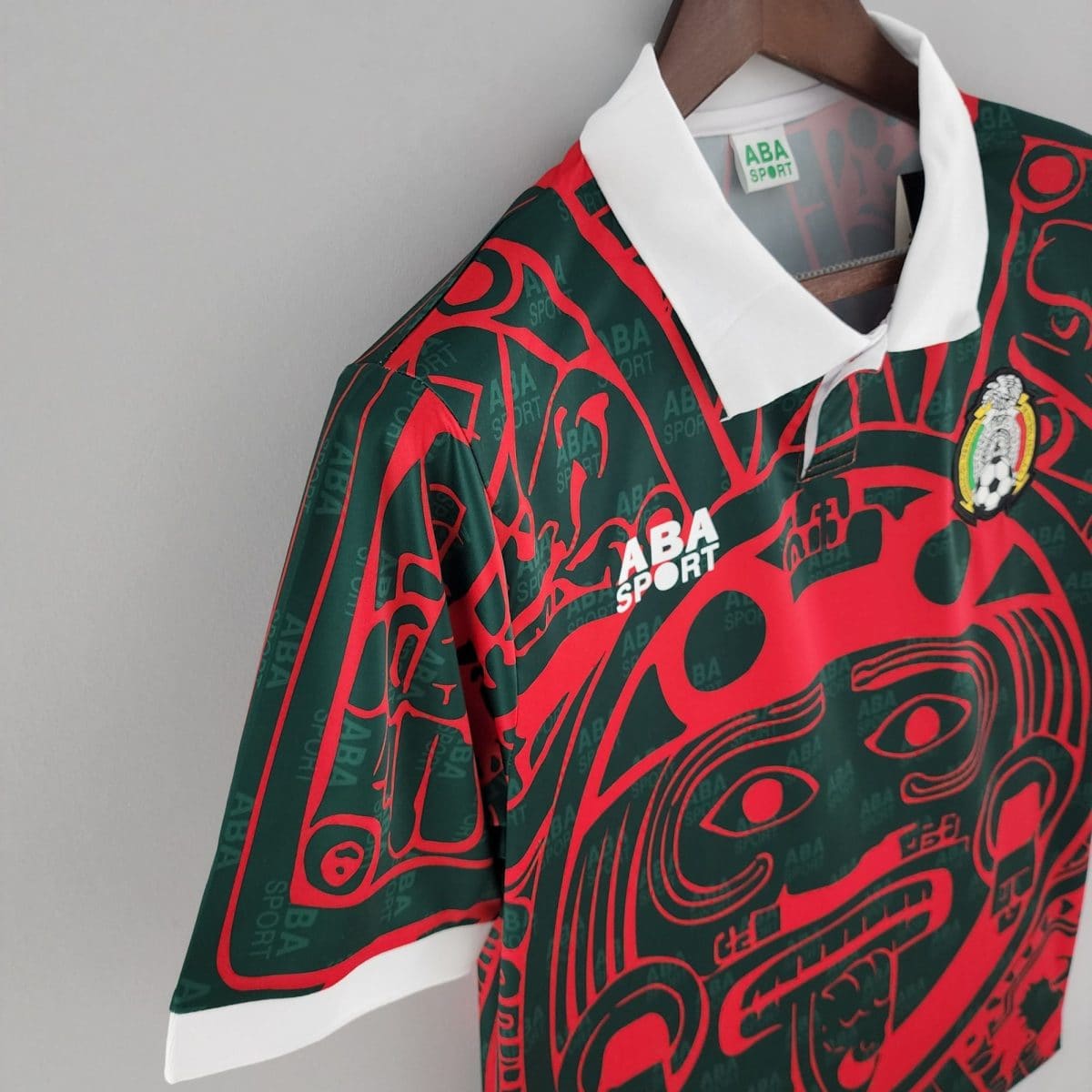 Close-up of a green and red patterned sports jersey on a wooden hanger with a white collar and visible ABA SPORT text in the pattern.