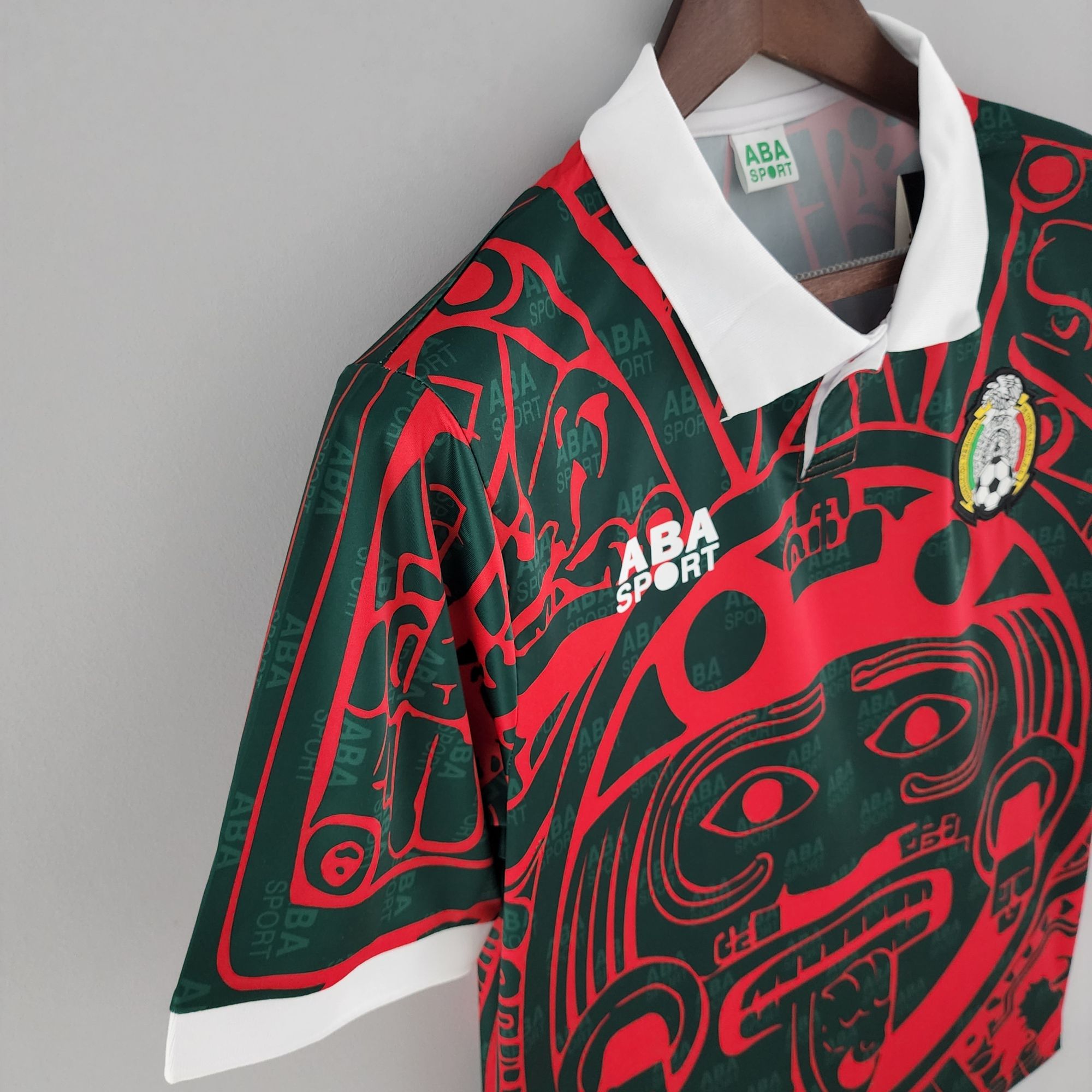 c10792e7_e7c738df-ea94-4230-9273-f26b9b7cbff4 | UAE SPORT STORE Close-up of a green and red patterned sports jersey on a wooden hanger with a white collar and visible ABA SPORT text in the pattern.