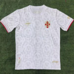 White football jersey with gold crest and subtle camouflage pattern, red trim at sleeve cuffs, laid on grass.