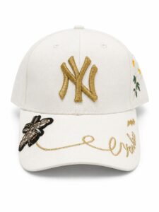 White baseball cap featuring a gold NY Yankees logo on the front, with floral embroidery on the side and gold cursive writing on the brim.