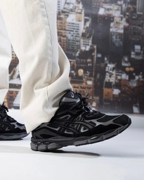 Close-up of black athletic sneakers stepping forward on a white surface, beige pants visible, with a blurred city skyline background.
