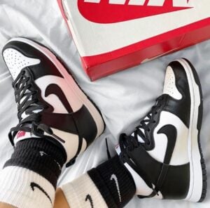 Two black-and-white Nike high-top sneakers worn with socks, resting near a red Nike shoebox on a bed.