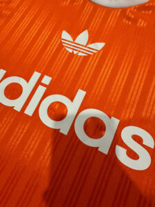 Close-up of an orange Adidas shirt showing the white trefoil logo and the letters Adidas on striped fabric.