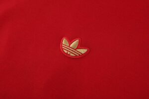Close-up of red fabric with a small gold Adidas trefoil logo embroidery.