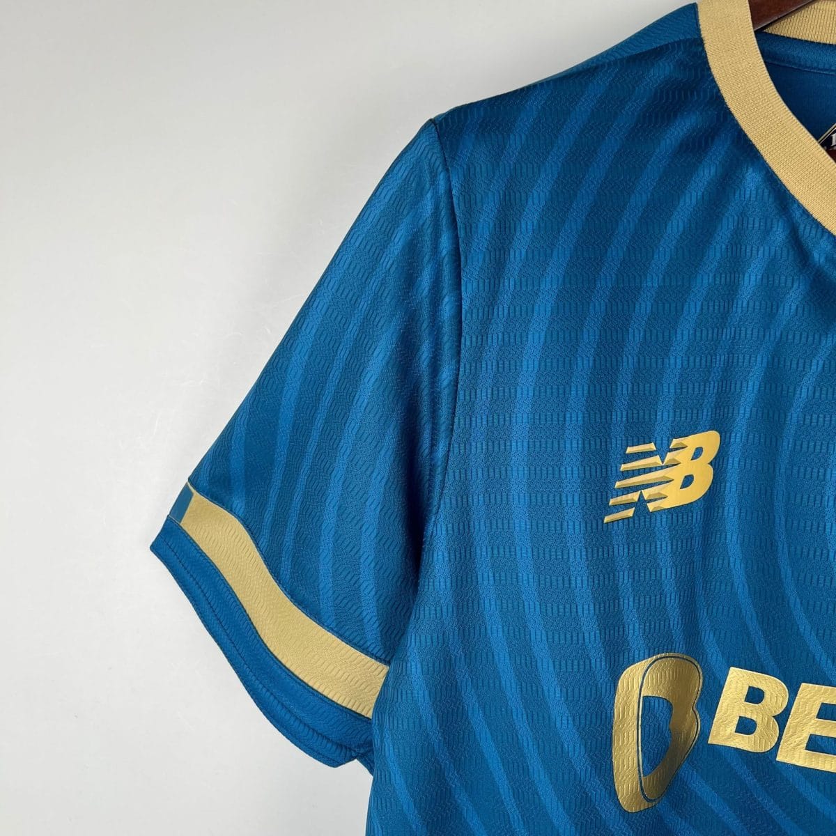 Close-up of a blue athletic jersey sleeve with a gold stripe and a partial gold New Balance logo badge nearby.
