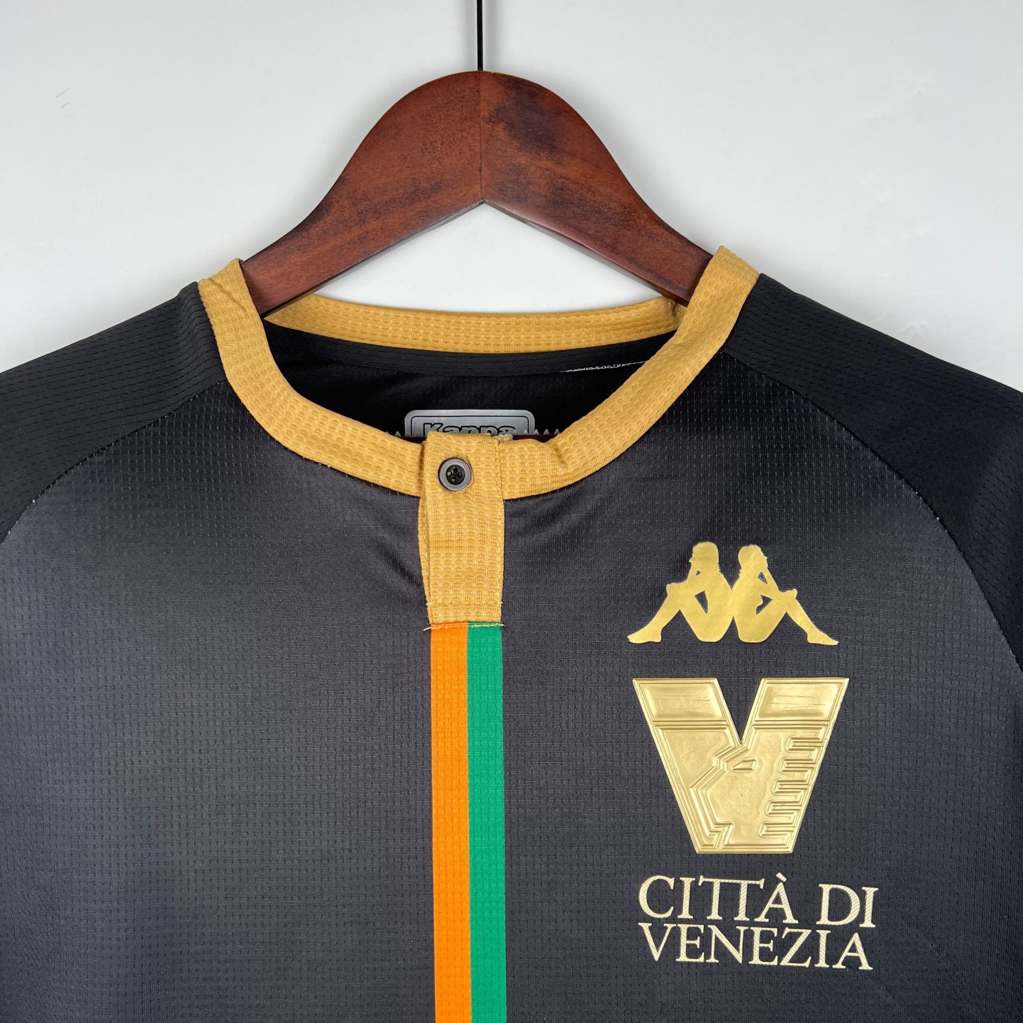 c347128f | UAE SPORT STORE قميص رياضي أسود بياقة صفراء وخط عمودي برتقالي-أخضر وشعار ذهبي على الصدر، معلّق على علاقة خشبية.