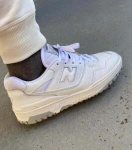 White New Balance 550 sneaker on a gray concrete floor, with a white sock and cuffed light pants visible, side view.