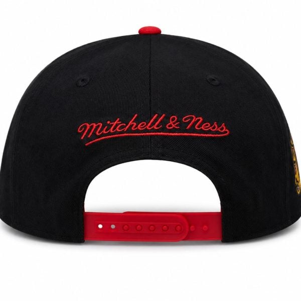 Back view of a black baseball cap with red embroidered 'Mitchell & Ness' lettering and a red adjustable snapback strap; white background.