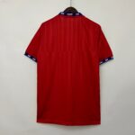 c3e138ae_a782ab40-dae8-4bdb-926b-3fb862ab1055 | UAE SPORT STORE Red short-sleeve striped shirt on a hanger, blue and white accented collar and cuffs, hanging against a light wall.