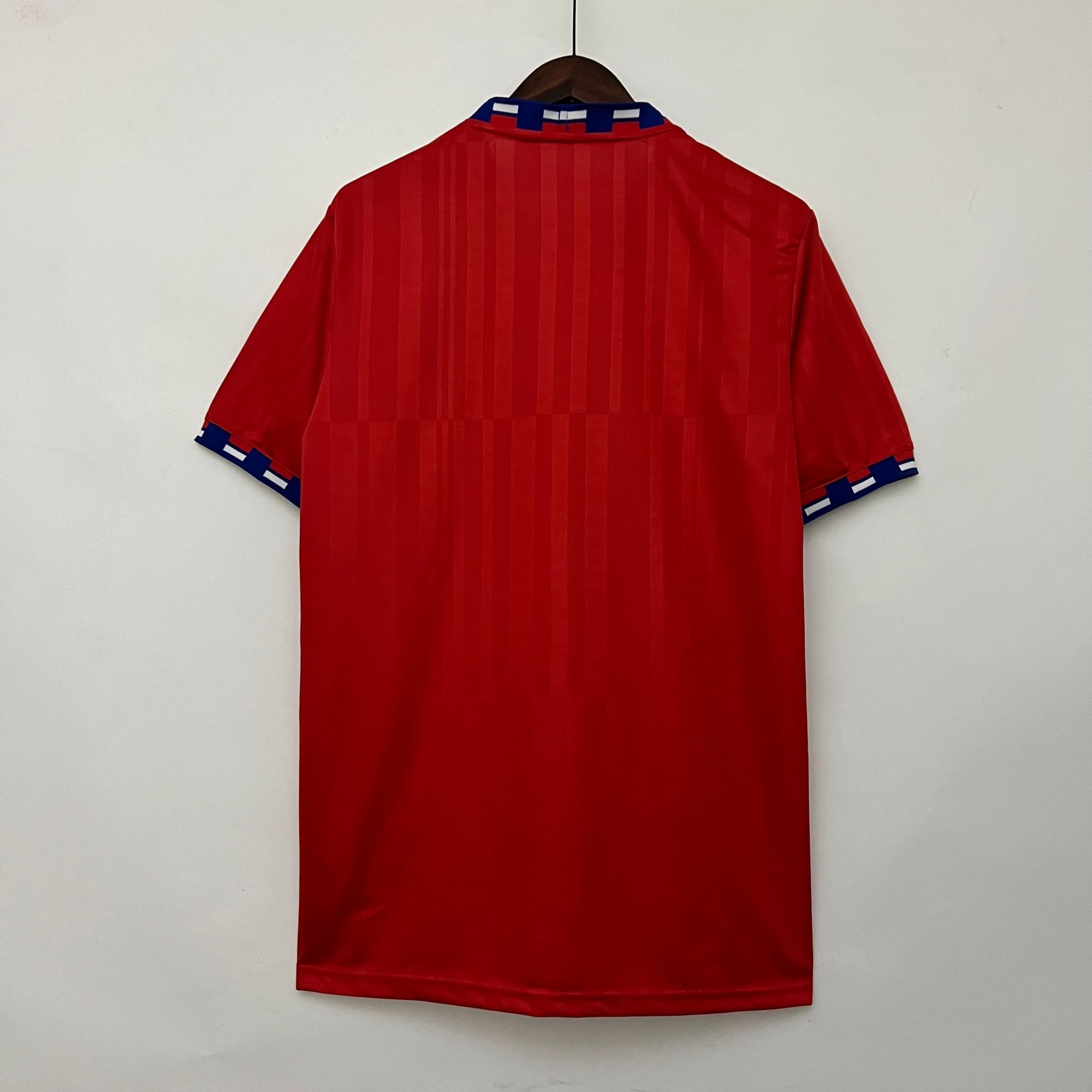 c3e138ae_a782ab40-dae8-4bdb-926b-3fb862ab1055 | UAE SPORT STORE Red short-sleeve striped shirt on a hanger, blue and white accented collar and cuffs, hanging against a light wall.