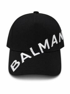 Black baseball cap with bold white embroidery spelling 'BALMAIN' diagonally across the brim.