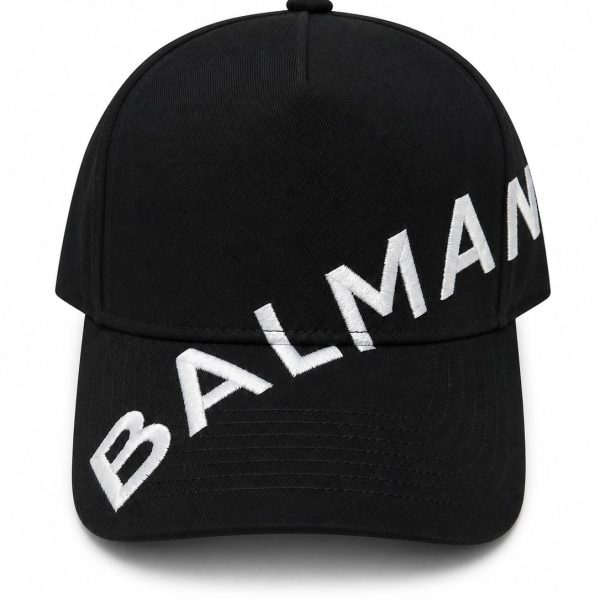Black baseball cap with bold white embroidery spelling 'BALMAIN' diagonally across the brim.