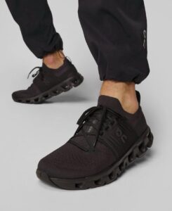 Black athletic sneakers with a bold, hollowed pod-like sole, worn by a person in black joggers stepping forward.