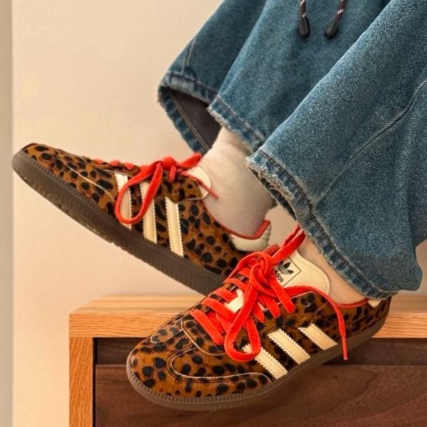 Leopard print sneakers with red laces and white stripes resting on a wooden surface, worn with jeans and white socks.