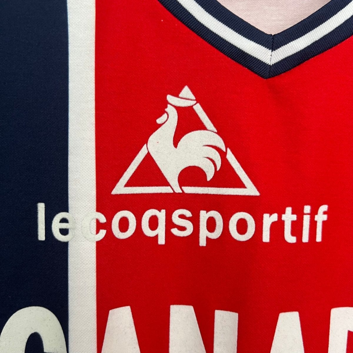 Close-up of red fabric showing part of the Le Coq Sportif logo — a white triangle with a white rooster silhouette and partial brand text visible on the red background.