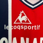 Close-up of red fabric showing part of the Le Coq Sportif logo — a white triangle with a white rooster silhouette and partial brand text visible on the red background.