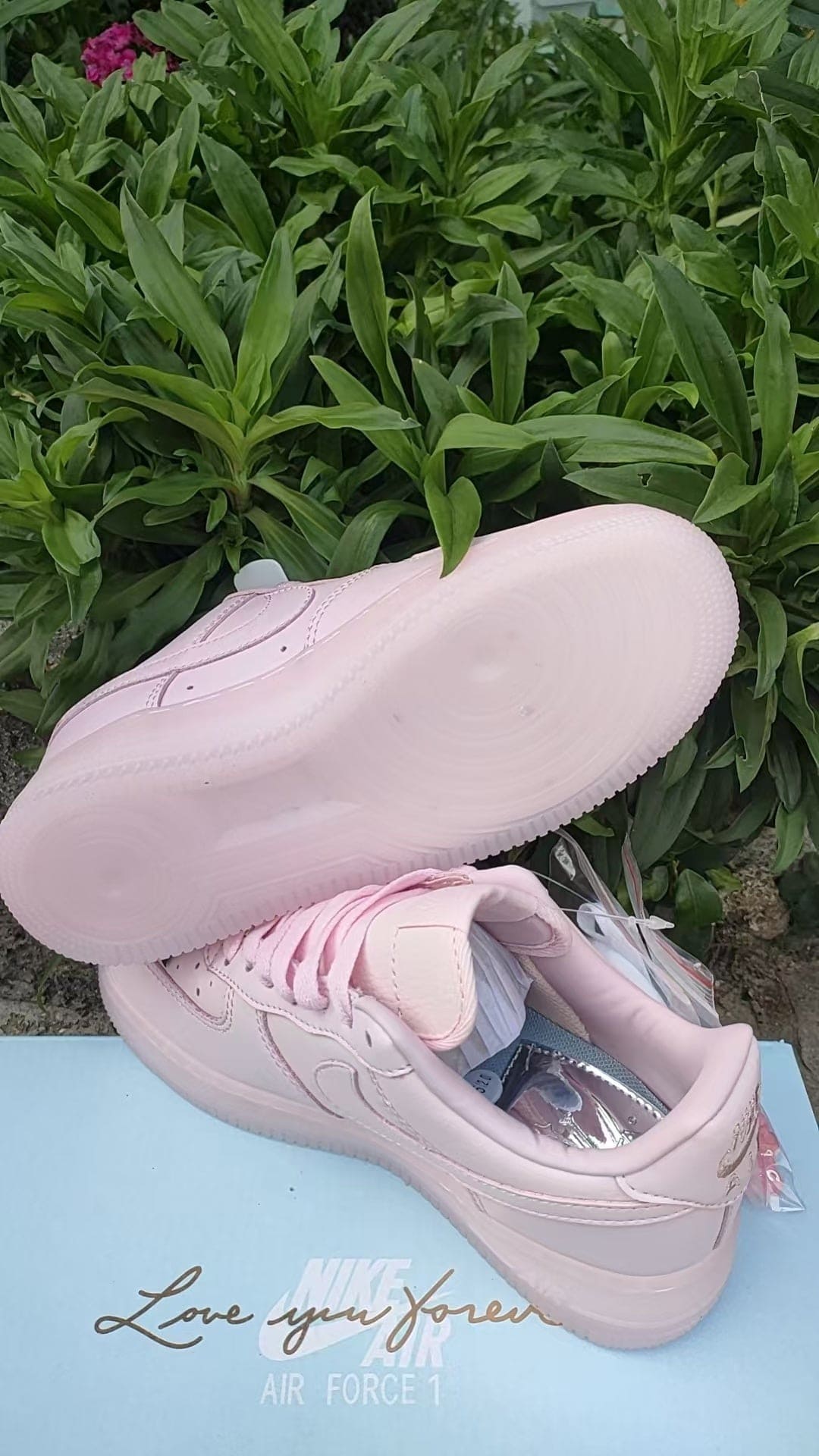 c487f475 | UAE SPORT STORE Pink Nike Air Force 1 sneaker resting on a light blue surface with green foliage in the background; text 'Love you forever' visible on the surface.