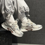 Close-up of white Nike sneakers on feet wearing white socks and light gray joggers, against a dark tiled wall.
