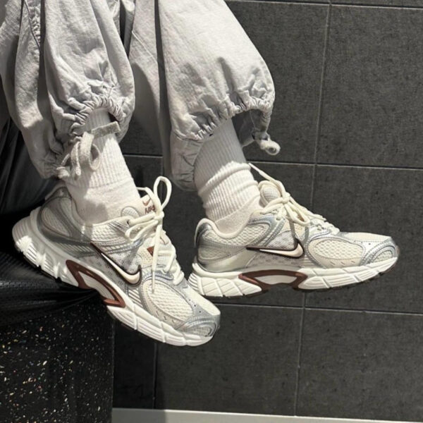 Close-up of white Nike sneakers on feet wearing white socks and light gray joggers, against a dark tiled wall.