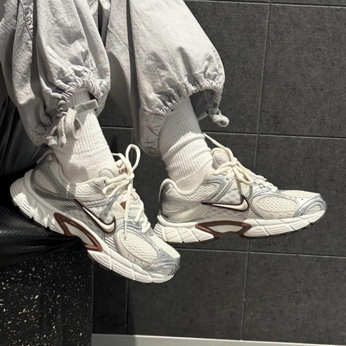c4aa9707-9f8c-4855-9dbf-3451e7cd198b Close-up of white Nike sneakers on feet wearing white socks and light gray joggers, against a dark tiled wall.