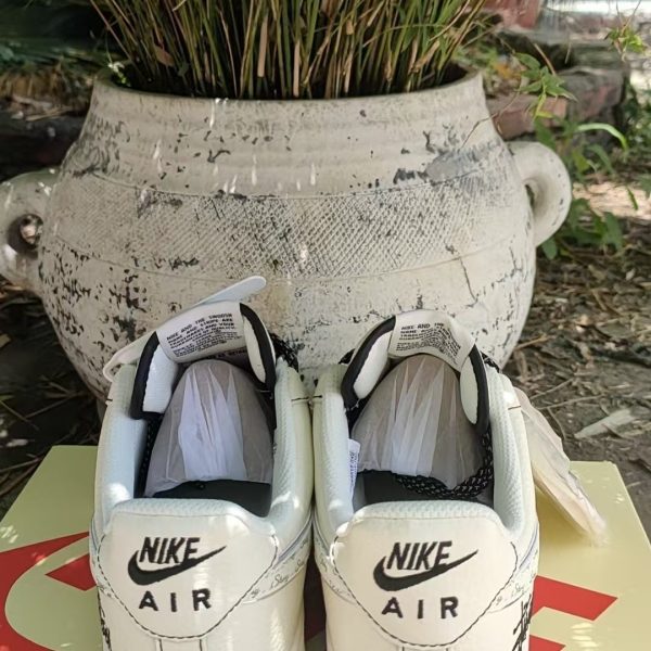 Pair of white Nike Air sneakers with black swoosh resting on a shoebox outdoors in front of a large white planter with green plants behind it.