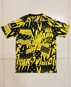 Bold yellow-and-black graphic T-shirt laid flat on a light tile floor, covered in chaotic typography and fleur-de-lis motifs.