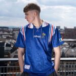 c4e83d52c130ef06547c07c1d732873a | UAE SPORT STORE Young man standing on a rooftop wearing a blue Adidas sports shirt with red and white stripes, looking to the left.
