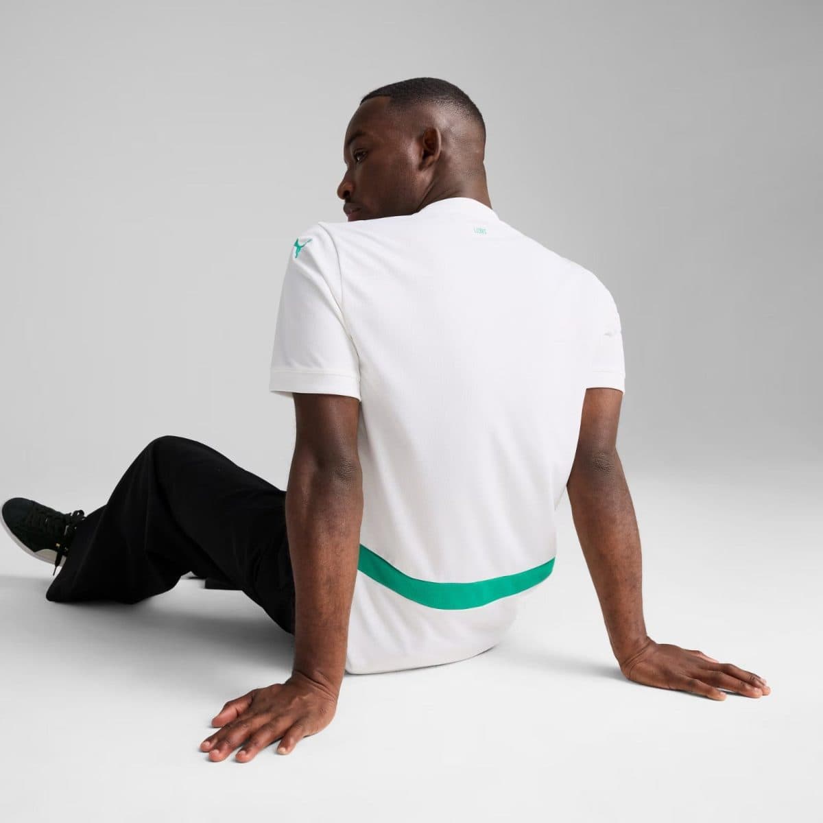 Back view of a man in a white polo with teal stripe around the waist, seated on the floor with hands flat behind him; small teal logo near the neckline.