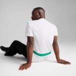 Back view of a man in a white polo with teal stripe around the waist, seated on the floor with hands flat behind him; small teal logo near the neckline.