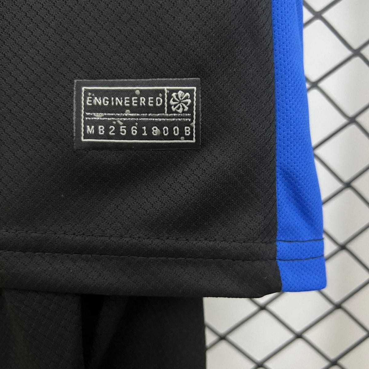 Close-up of black fabric with a stitched label reading 'ENGINEERED' and a small emblem, blue strap on the right, with a chain-link fence in the background.