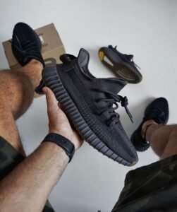 Person holding a black knit sneaker with a chunky ridged sole; another shoe and a shoebox are on the floor in the background.