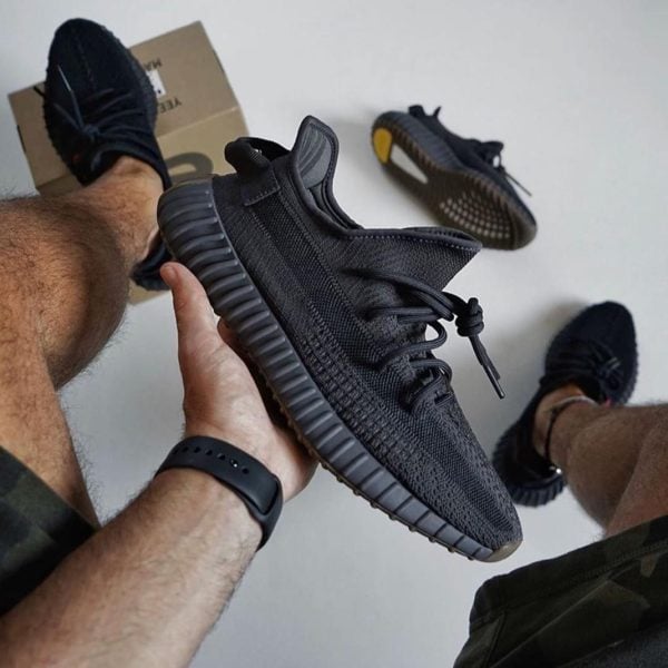 Person holding a black knit sneaker with a chunky ridged sole; another shoe and a shoebox are on the floor in the background.