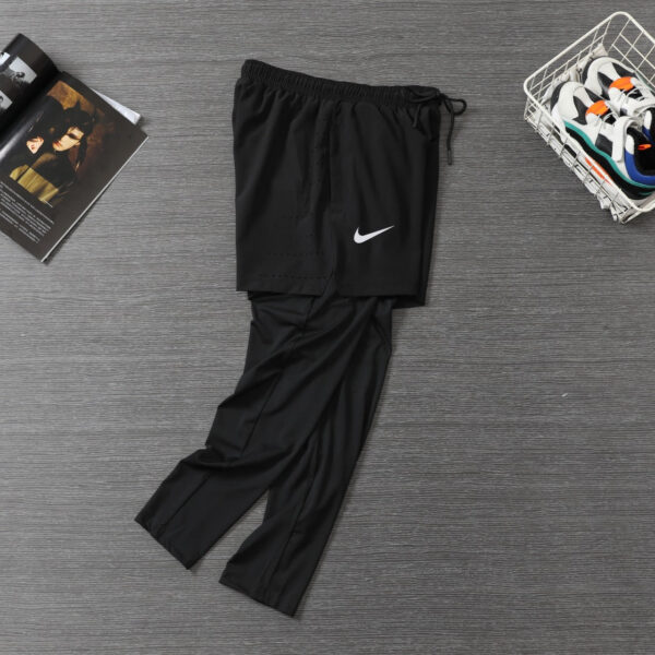 Black Nike athletic pants laid flat on a wooden floor with a white swoosh logo, a sneaker basket to the right, and a magazine nearby.
