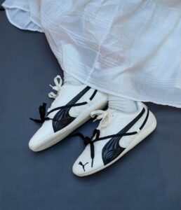 c789b311-de51-43cd-bdaa-f40b5001d64f | UAE SPORT STORE White sneakers with black stripes and laces on a dark floor, worn with white socks. The shoes feature a small black logo on the toe.