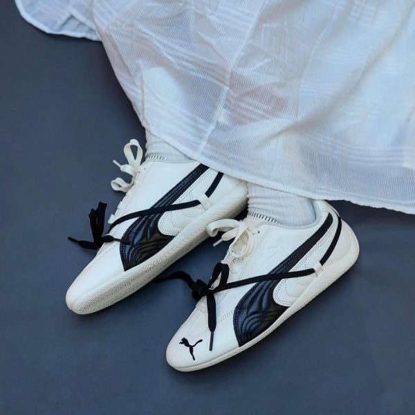 White sneakers with black stripes and laces on a dark floor, worn with white socks. The shoes feature a small black logo on the toe.
