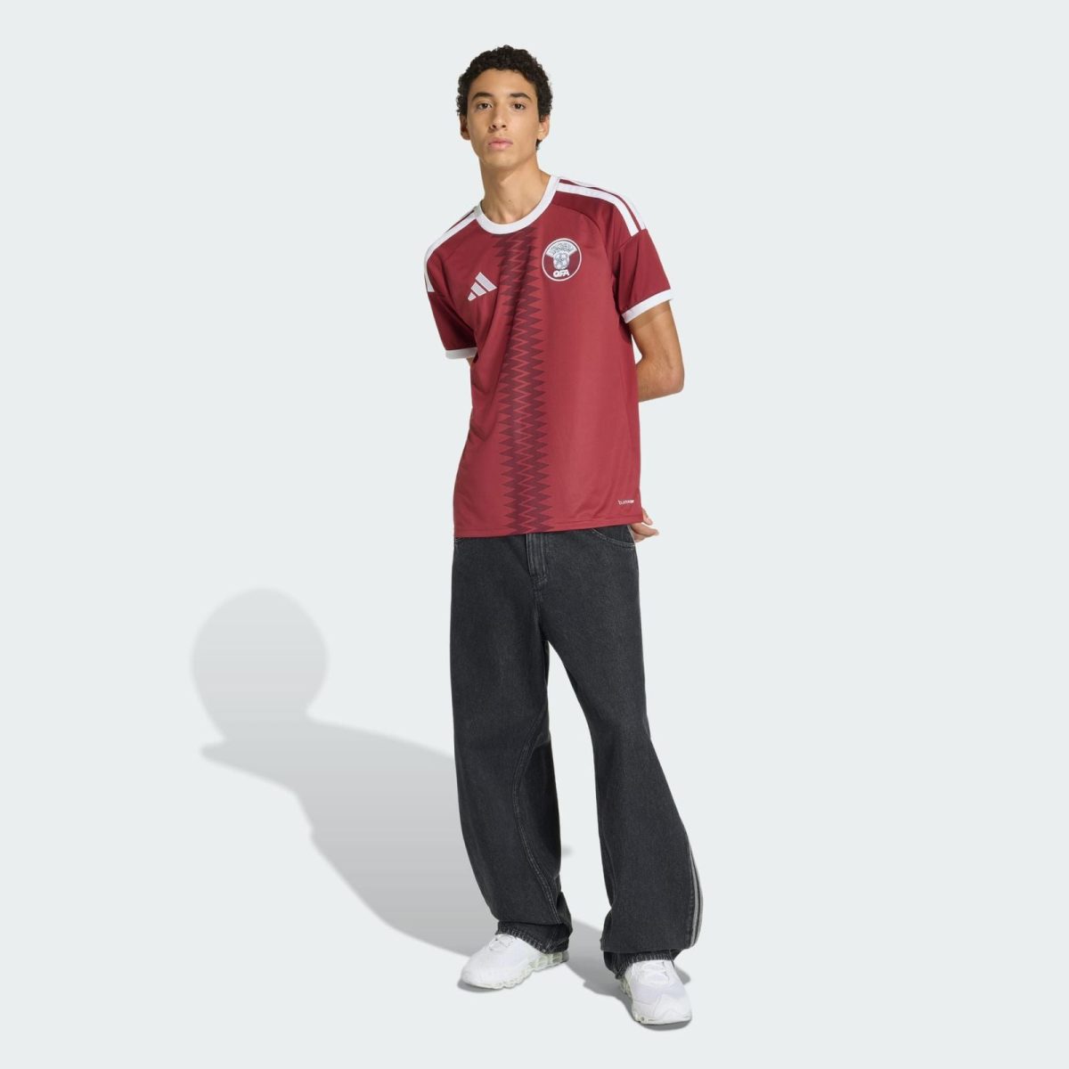 Person wearing a maroon Adidas soccer jersey with geometric stripe, dark jeans, and white sneakers, standing against a pale background.