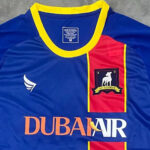 Blue and yellow soccer jersey with a red panel and team crest on the chest; sponsor text 'DUBAI AIR' across the front.