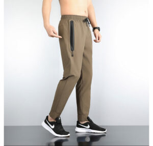 Side view of a man modeling tan jogger pants with a vertical zip pocket on the thigh and cuffed ankles, wearing black sneakers.