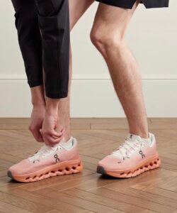 Person bending down to tie the laces of pink-orange athletic shoes on a wooden floor.